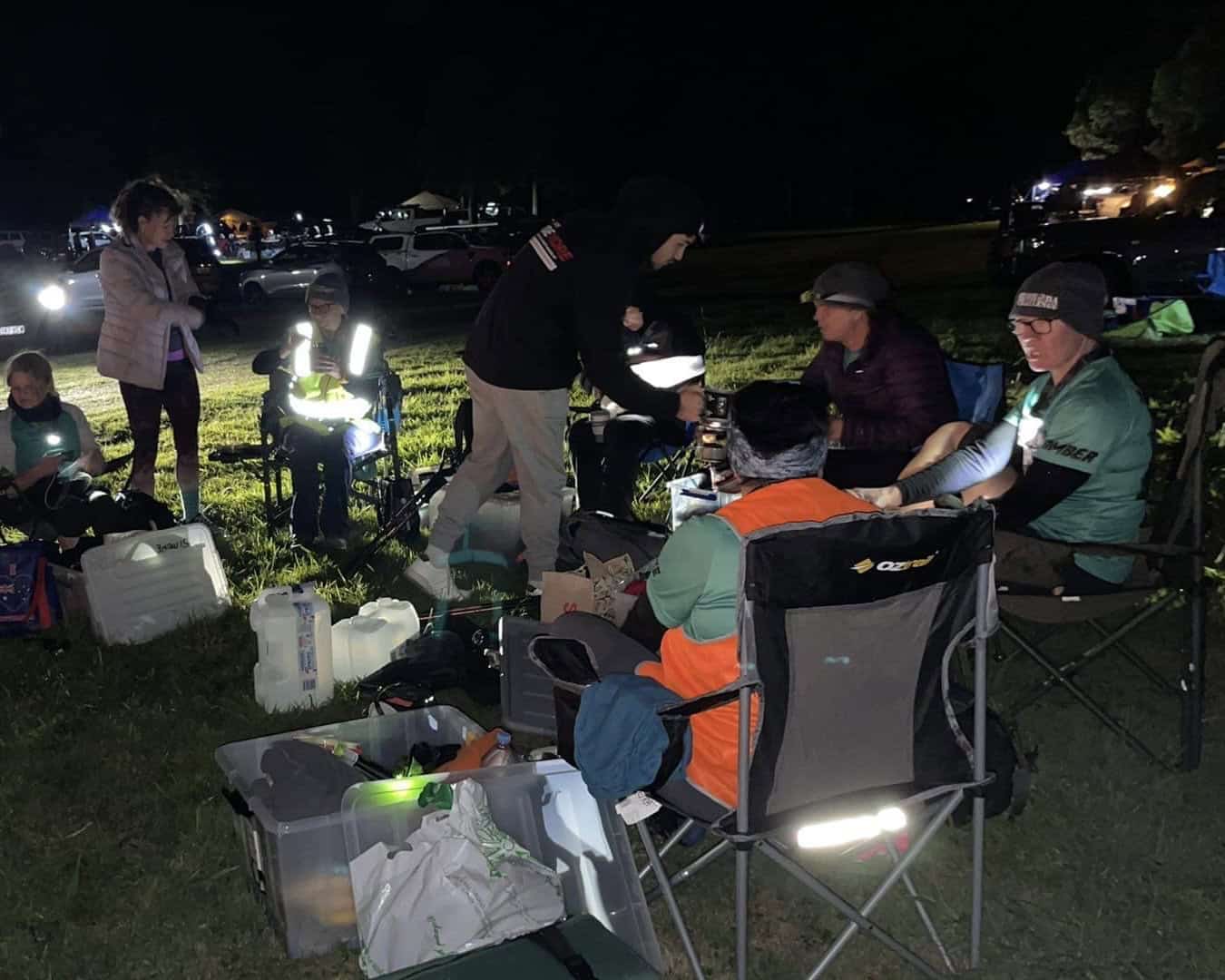 A group of people, likely part of the Kokoda Challenge support crews, sit and stand around camping gear and boxes at night, illuminated by flashlights and headlamps in a grassy area. Some are wearing reflective vests.