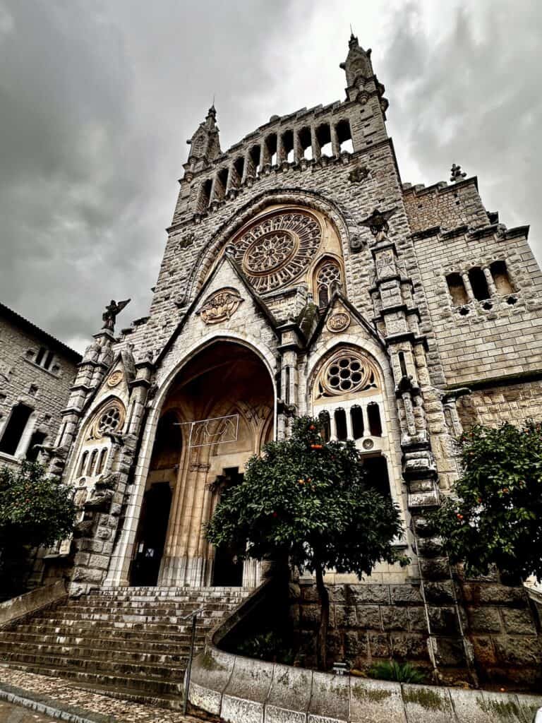 A gothic-style cathedral with a detailed facade and large archway, flanked by two orange trees. The sky is overcast.