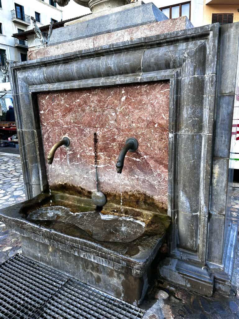 A stone fountain with two water spouts set in a red marble panel. Water is flowing from both spouts into a rectangular basin. The surrounding area is paved with stone tiles.