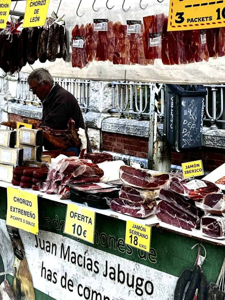 A vendor stands behind a market stall displaying various types of cured meats, including chorizo and jamón. Signs indicate prices and offers for different products.