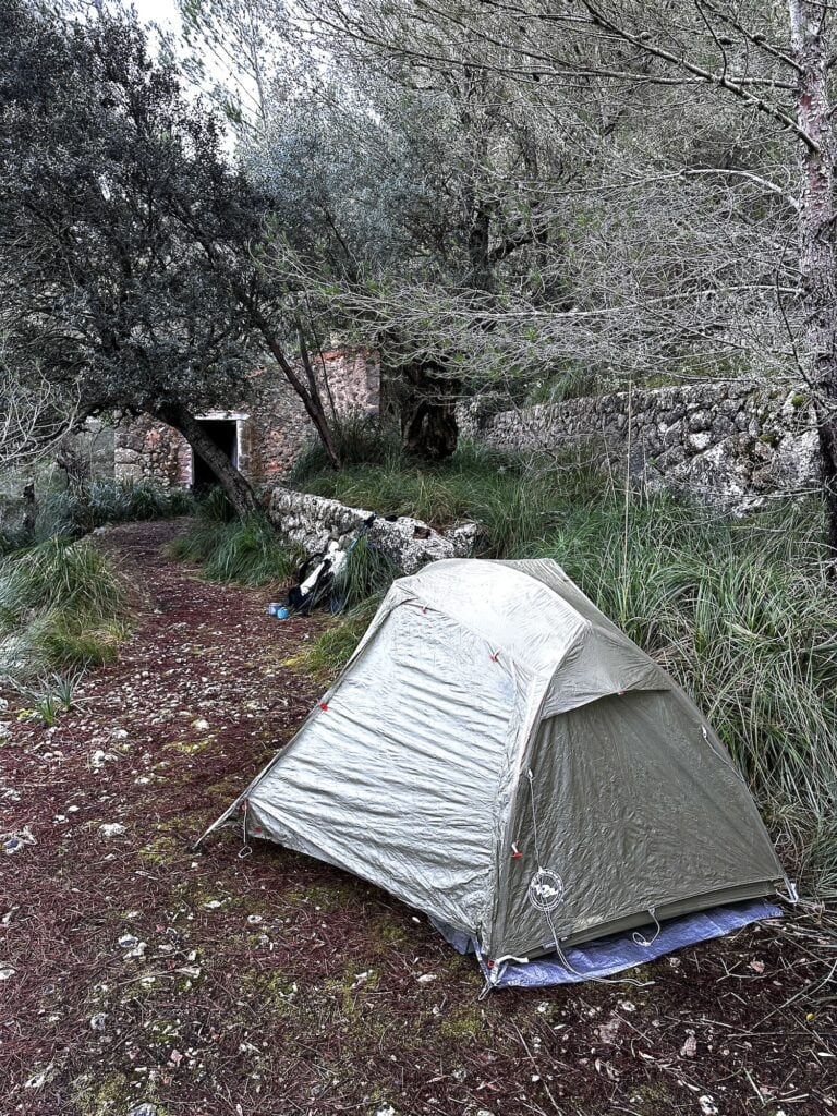 A tent is pitched on a forest path next to an old stone building. The scenery is lush, with trees and grass surrounding the area. Camping gear is placed outside the tent.