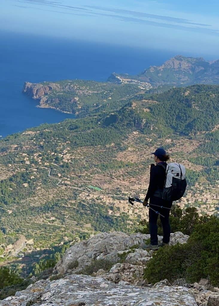A person with a backpack and trekking poles stands on a rocky edge overlooking the vast landscape of hills, villages, and the ocean in the distance, while traversing the renowned GR221 dry stone route.