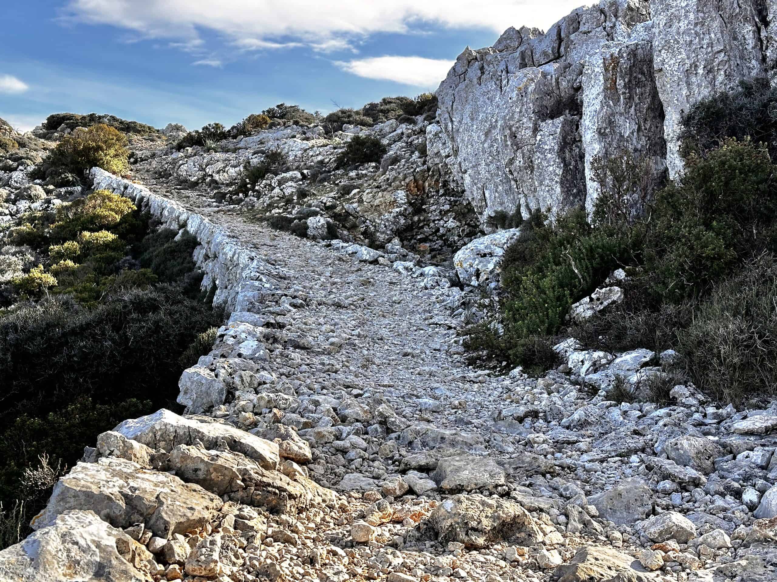 A rocky path winds through a rugged, mountainous landscape with large boulders and sparse vegetation under a blue sky, marking a scenic stretch of the GR221 dry stone route in Mallorca.