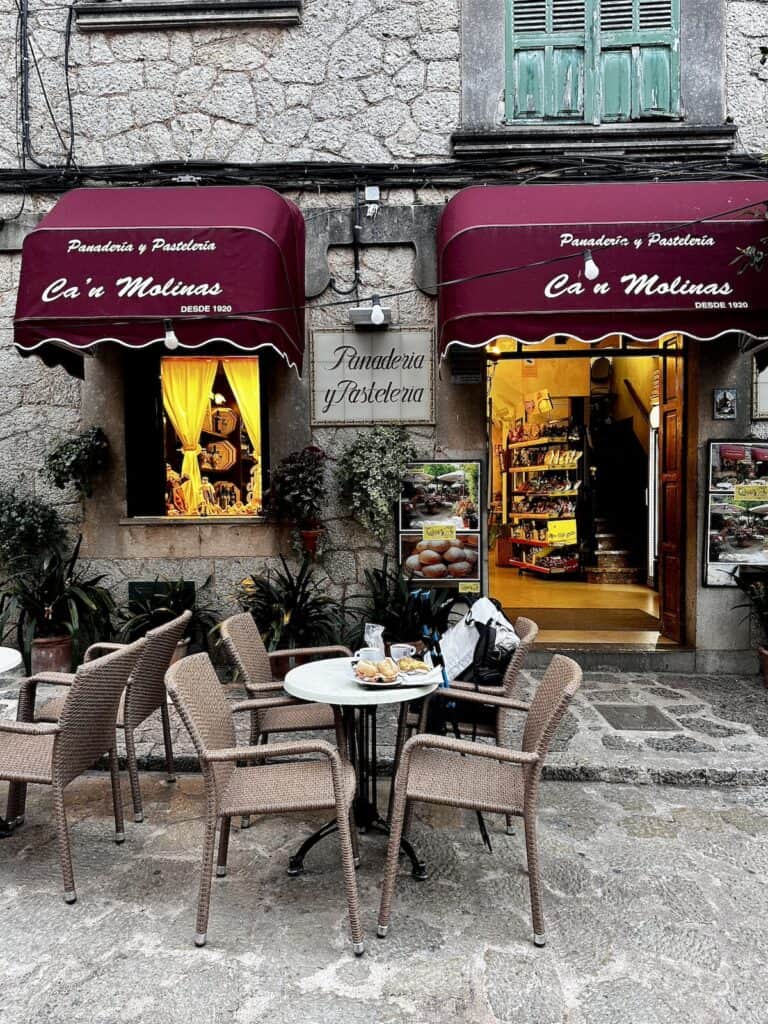 Outdoor seating area of a bakery and patisserie with maroon awnings, wicker chairs, and stone walls reminiscent of the GR221 dry stone route in Mallorca. The storefront has an open door and window displaying pastries.