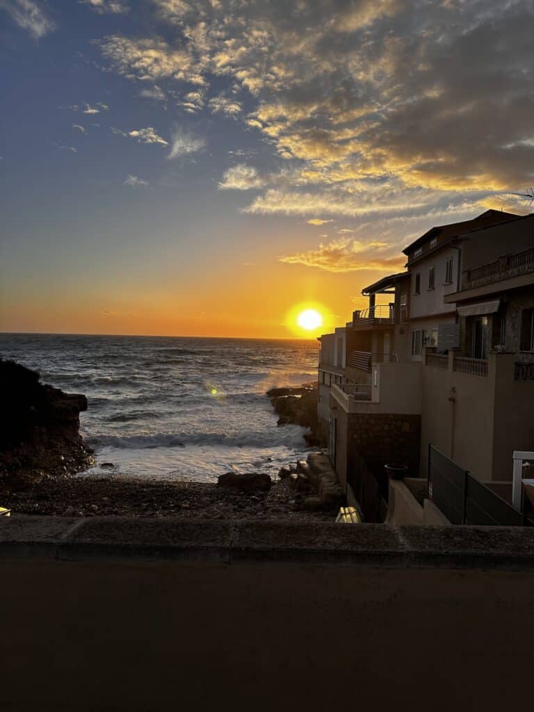 A sunset over a coastal view with waves crashing onto a rocky shore and houses on the right side. The sky has scattered clouds with hues of orange and yellow as the sun sets on the horizon.
