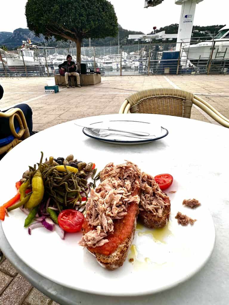 Plate with bread topped with tuna, oil, and tomato alongside mixed vegetables and olives on an outdoor table by a waterfront with boats and seated people in the background, a perfect stop while exploring the GR221 Mallorca dry stone route.