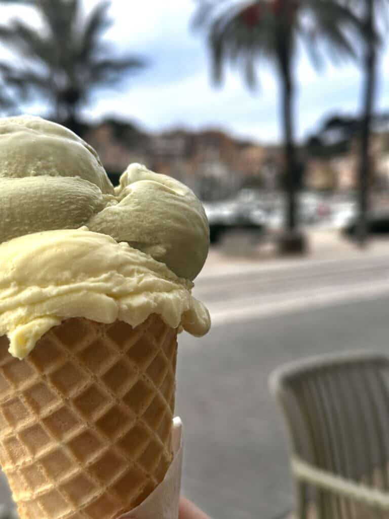 Close-up of an ice cream cone with two scoops, vanilla and pistachio, held in hand. Background shows blurred palm trees and buildings.