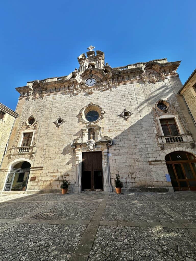 A historic stone building with intricate carvings, a central entrance, three arched windows, and a clock tower under a clear blue sky.