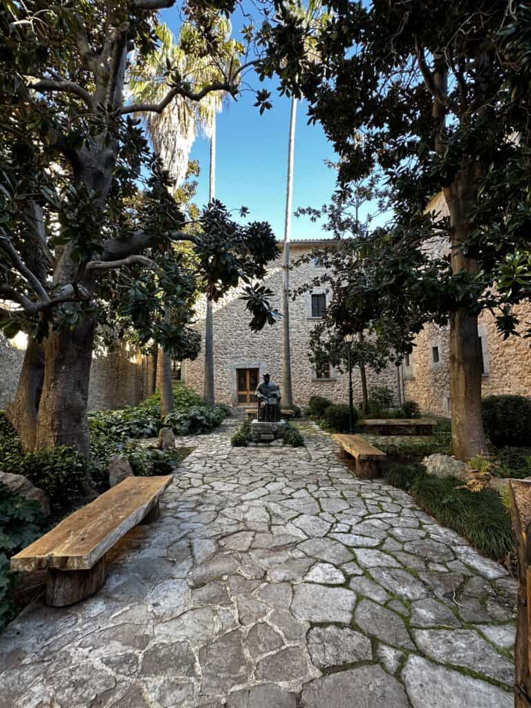 Stone courtyard with tall trees, benches, and a central statue. The setting is surrounded by stone buildings, and the sky above is clear and blue.