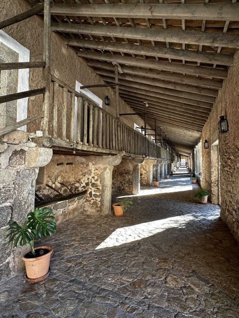 A long, covered stone corridor with wooden railings, potted plants along the walkway, and sunlight streaming through the gaps.