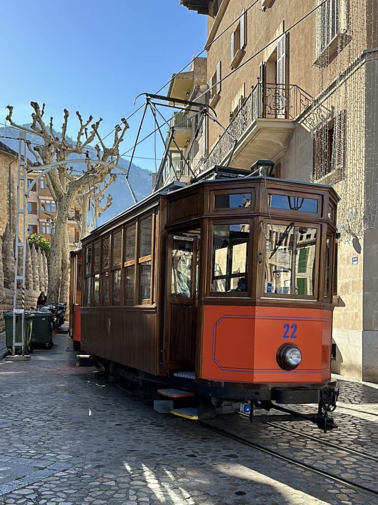 A vintage orange tram with wooden panels and the number 22 on the front is stationed on a cobblestone street in a town with leafless trees and traditional buildings.
