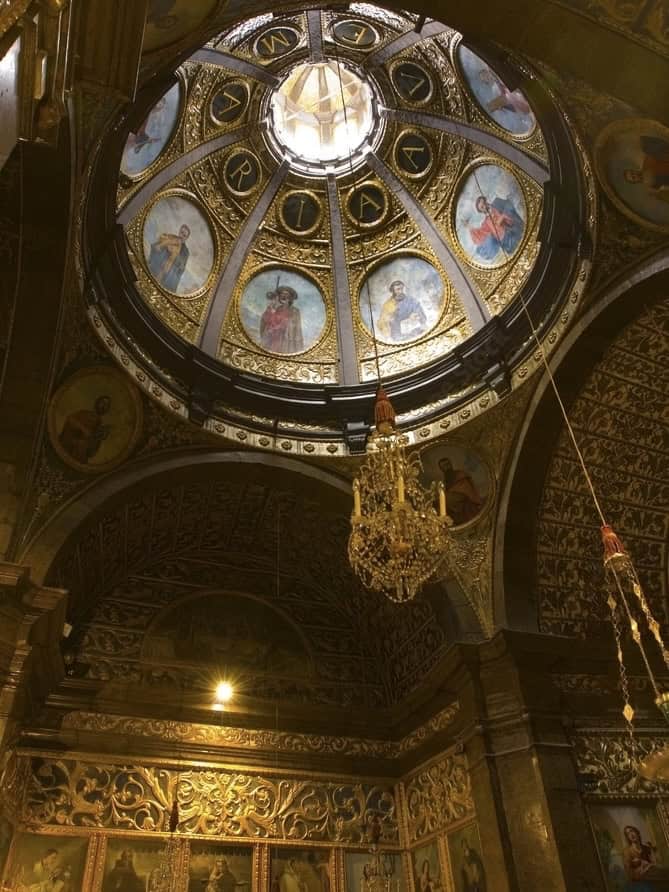 Ornate domed ceiling of a church featuring circular paintings of saints, intricate gold designs, and a central chandelier.