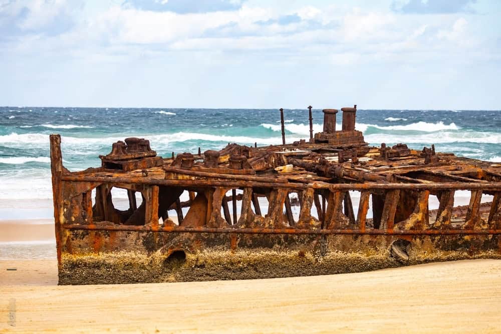 Rusty, partially submerged shipwreck on a sandy beach with waves in the background under a cloudy sky, serving as a reminder to follow leave no trace principles.