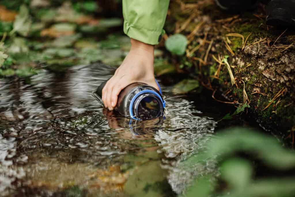 A person's hand filling a glass jar with water from a small stream, surrounded by green vegetation, following tips for hiking in hot weather.
