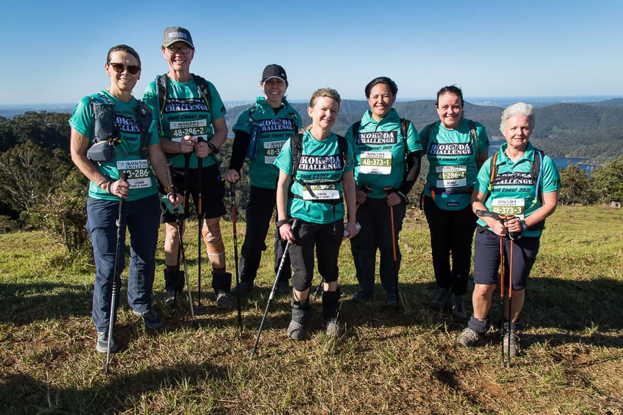A group of seven people wearing matching green shirts and holding trekking poles stand on a grassy hilltop with hills and a body of water in the background, embodying the spirit of the Kokoda Challenge support crews.