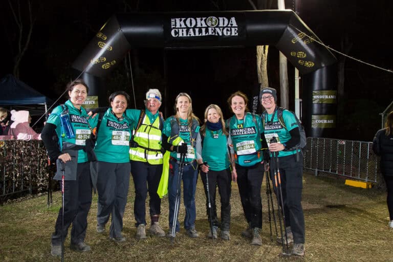 A group of people in teal shirts stands together with trekking poles under an archway that says "The Kokoda Challenge." They appear to be part of an event or competition, likely with the kokoda challenge support crew backing their efforts.