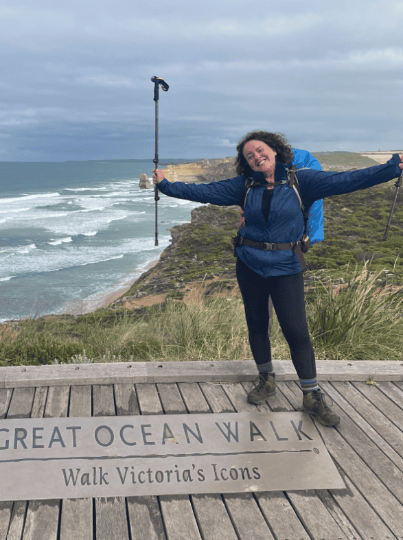 A person stands with arms outstretched and hiking poles in hand on a wooden platform overlooking a coastal landscape with the sign "Great Ocean Walk: Walk Victoria's Icons" visible on the ground.