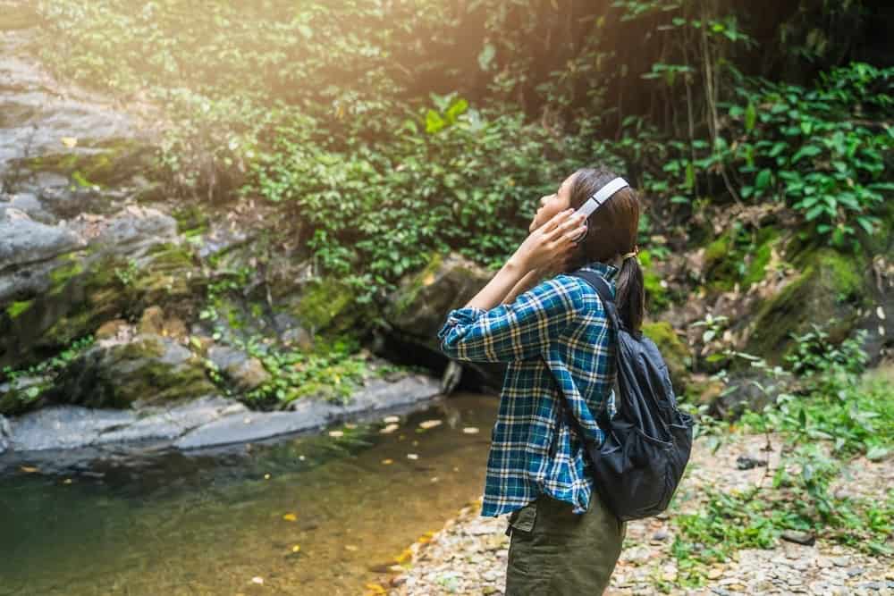 A person wearing a plaid shirt and headphones stands by a rocky creek in a lush forest, looking upwards with hands on the headset, embodying the leave no trace principles to preserve the natural beauty around them.