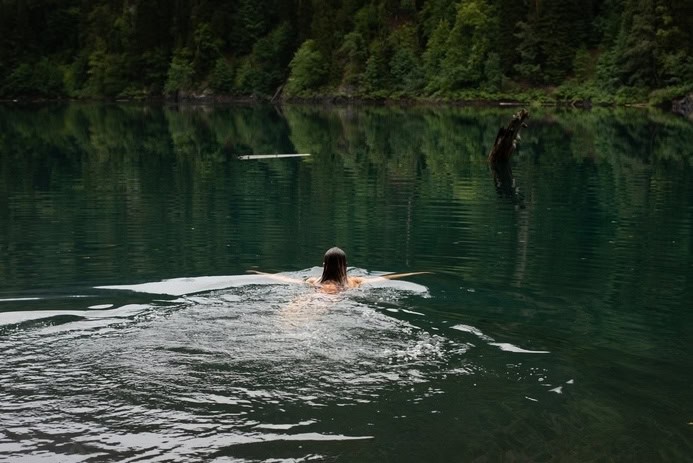 A person swims in a calm lake surrounded by dense forest, mindful of the principles of Leave No Trace to protect this serene wilderness.