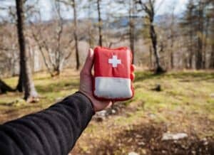 A person holds a red and white hiking first aid kit in a wooded outdoor area.