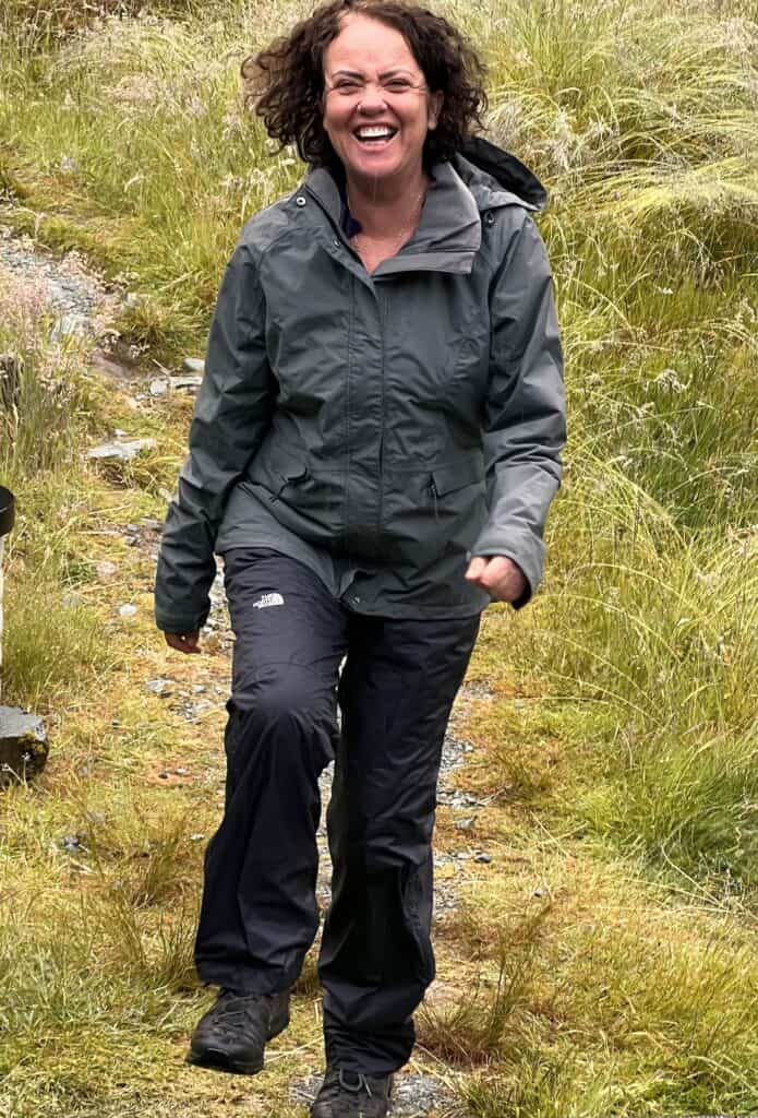 A woman in outdoor attire, including a gray jacket and black pants, is walking on a grassy path, smiling widely.