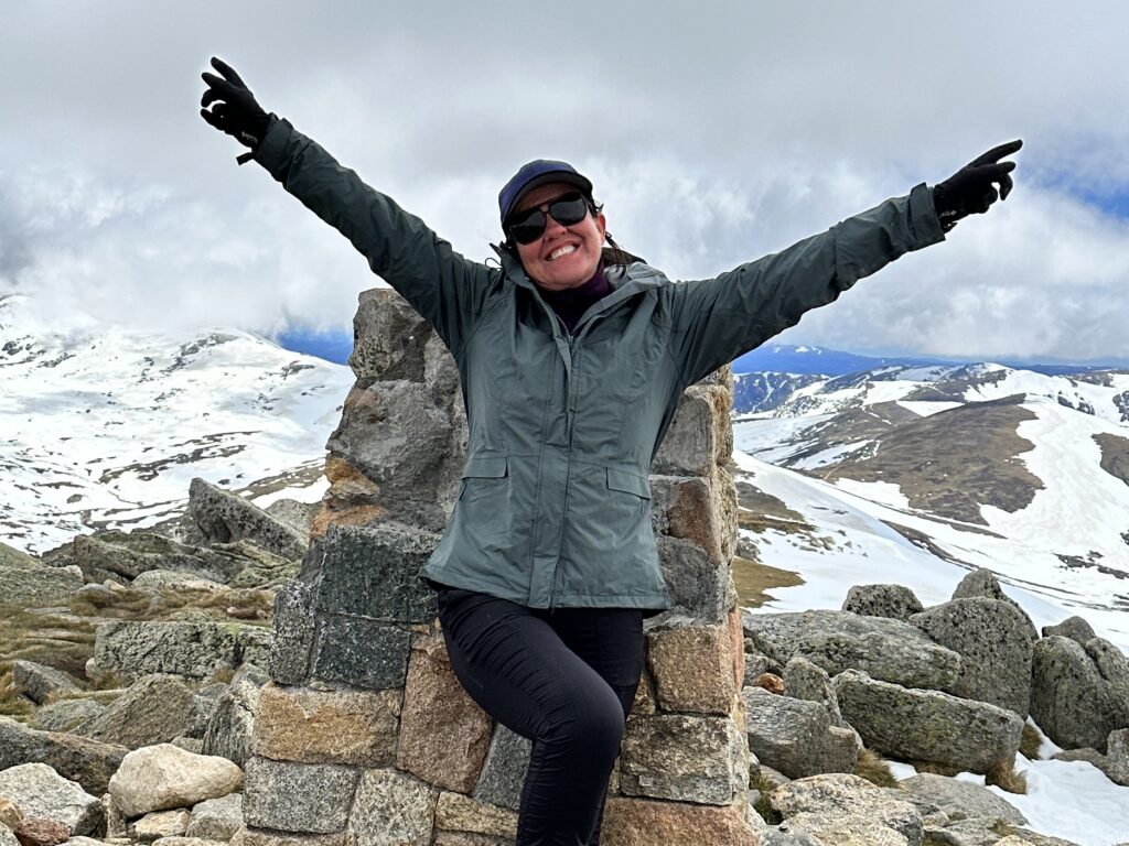 woman standing against a cairn on top of a mountain with hr arms spread out 
