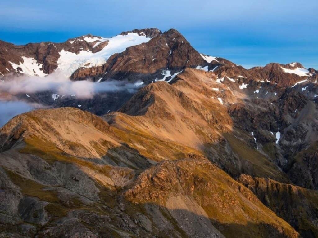 A stunning mountain range with rugged peaks, partially covered in snow and ice, is bathed in warm sunlight. Low clouds float around the mountains, while the sky above is clear and blue, creating a serene and majestic landscape.