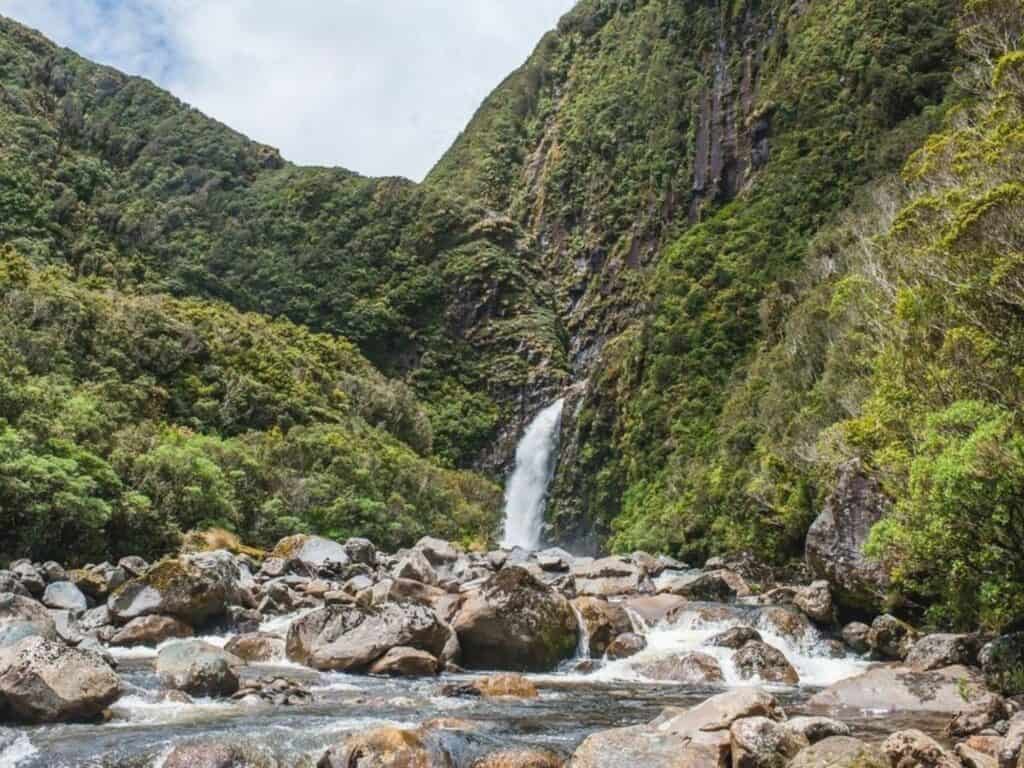 A waterfall cascades down a lush green mountainside, surrounded by dense foliage. Rocks and small boulders line the foreground, with a clear stream flowing over them under a partly cloudy sky—an ideal spot for hiking in North Island New Zealand's breathtaking landscapes.