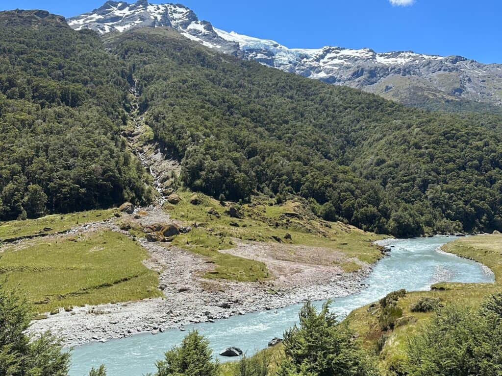 A winding river flows through a lush green valley with dense forests, leading to snow-capped mountains under a bright blue sky. The sunlight highlights the vibrant landscape and contrasts with the white peaks in the background.