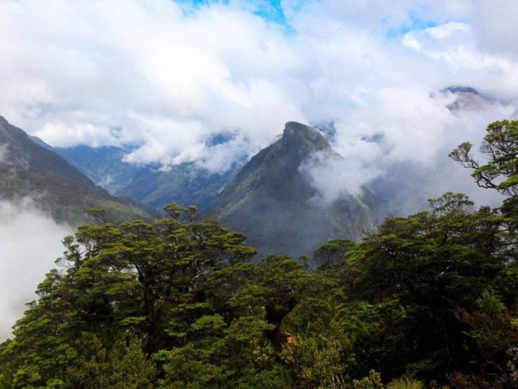 A lush mountain landscape with mist-covered peaks and dense greenery in the foreground. Clouds drift around the mountains, partially obscuring them, creating a serene and mystical atmosphere under a blue sky.