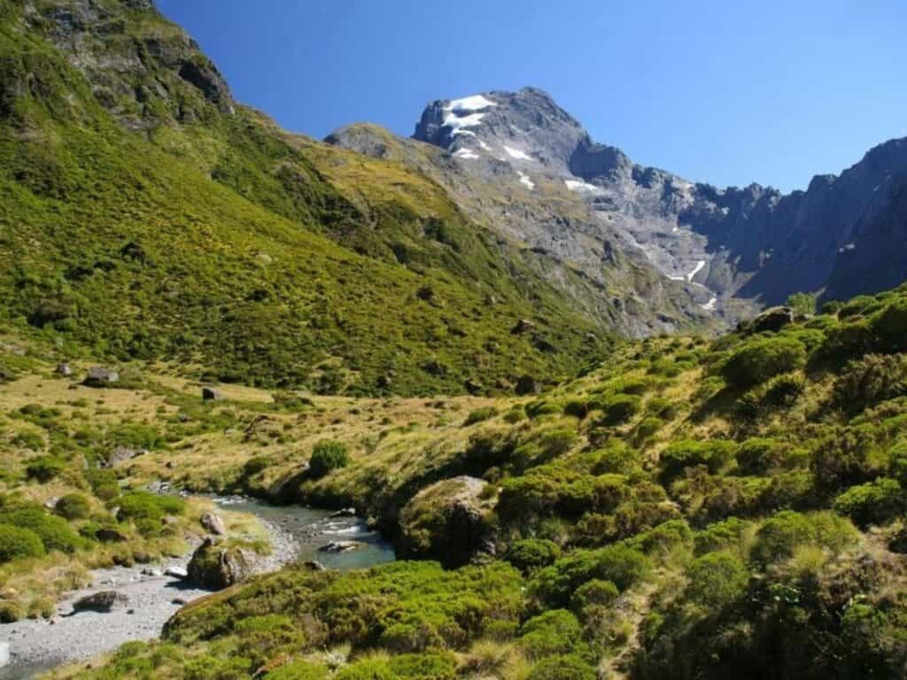 A scenic view of a mountain landscape with a rocky peak partially covered in snow. The foreground features a lush green valley with small hills and a winding stream under a clear blue sky.