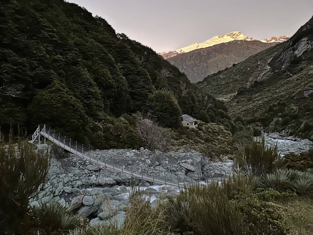 A suspension bridge crosses a rocky stream in a lush, green valley. Dense trees cover the hills, leading up to a snow-capped mountain peak illuminated by the sunset. A small cabin sits nestled among the foliage.