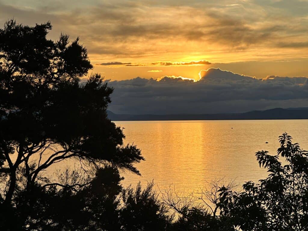 A serene sunset over a calm body of water, with the sun partially obscured by clouds, evokes the tranquil end to a day of hiking New Zealand's North Island. The sky is painted in shades of orange and gold, reflecting on the water as silhouettes of trees frame this picturesque scene.