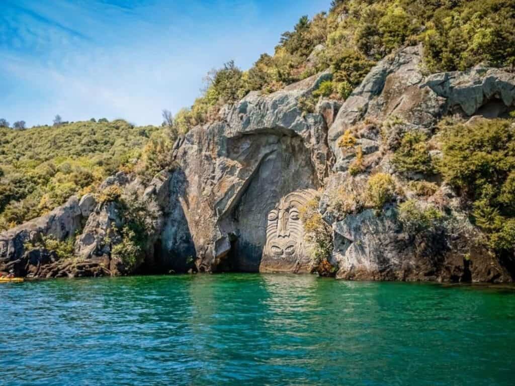 Nestled in New Zealand's North Island, this serene lake boasts a large rock carving of a face set into a rocky cliff, surrounded by lush vegetation under a clear blue sky. The turquoise water gently reflects the natural scenery, offering a tranquil pause on your hiking adventure.