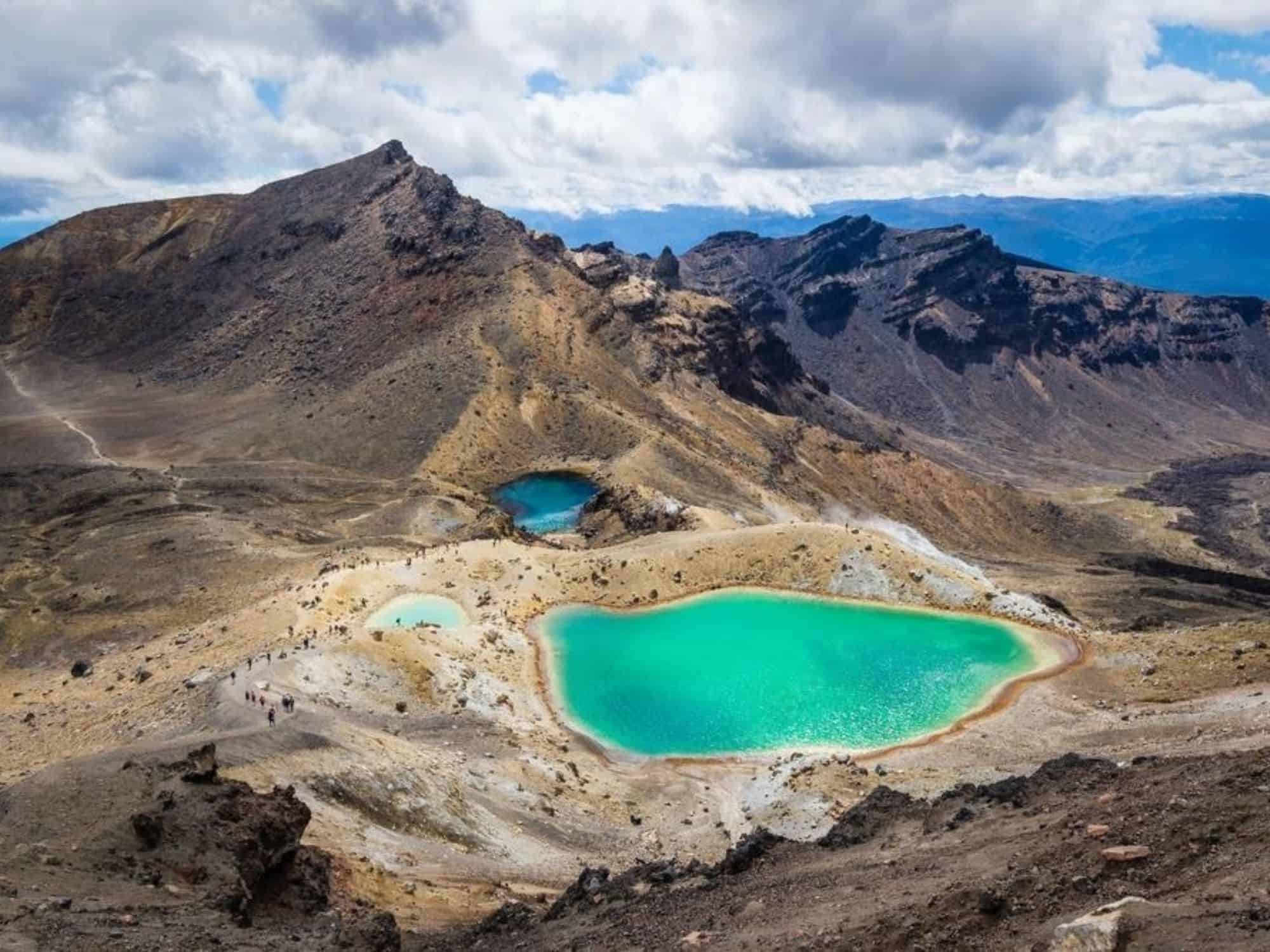 Two vibrant turquoise lakes amidst a rugged, rocky landscape on New Zealand's North Island. The lakes are surrounded by barren, brown terrain with distant mountains under a partly cloudy sky. Small figures of hikers are visible on a trail near the lakes, drawing nature enthusiasts from afar.