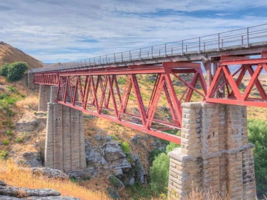 A red steel truss bridge spans across a rocky landscape with grassy hills and shrubs. The bridge is supported by stone pillars, and a blue sky with scattered clouds is overhead.