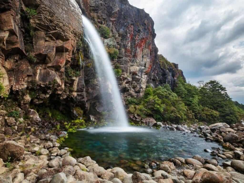 A waterfall cascades down a rocky cliff into a clear pool, surrounded by rocks and lush green foliage, making it an ideal spot for hiking New Zealand's North Island. The cloudy sky adds a dramatic backdrop to the serene landscape near Mount Taranaki.