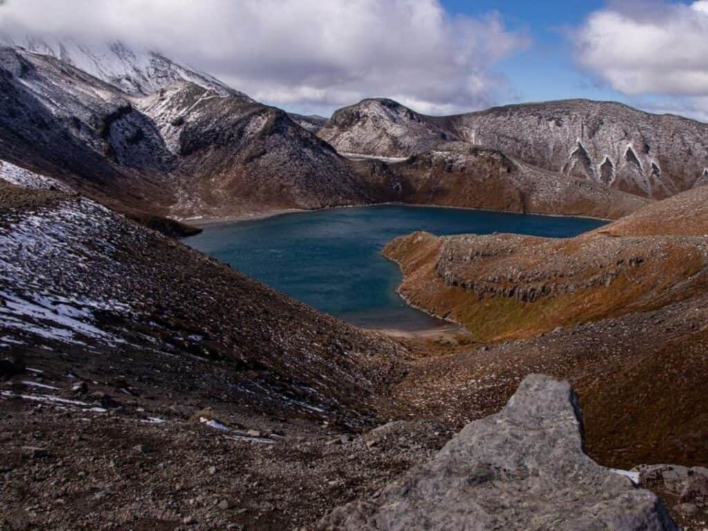 A serene, blue lake on New Zealand's North Island is surrounded by rugged, snow-dusted mountains under a partly cloudy sky. The landscape, perfect for hiking Mount Taranaki, features rocky terrain and patches of snow, creating a tranquil and isolated natural scene.