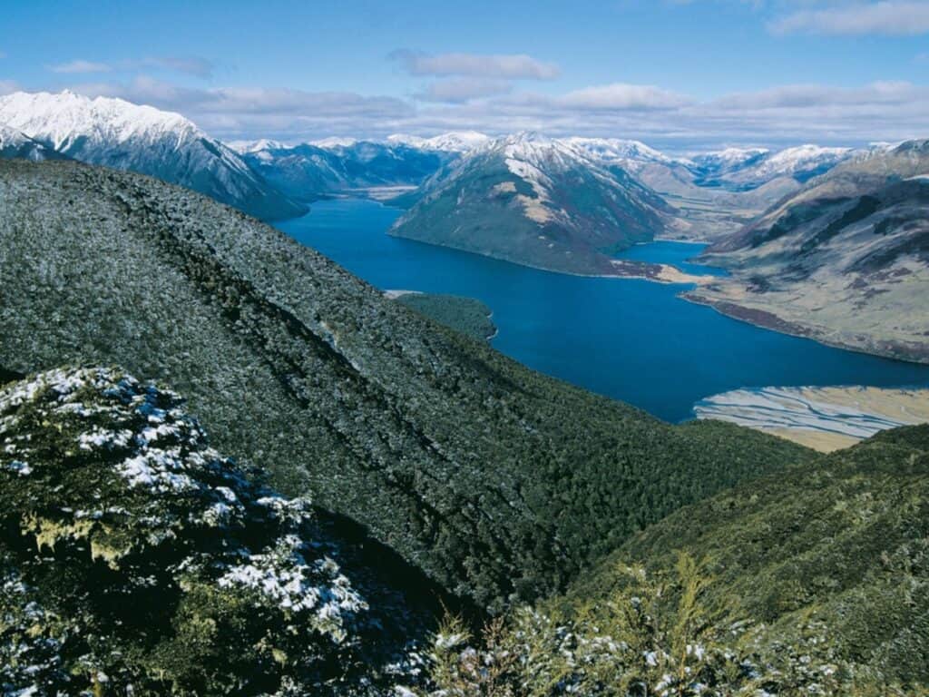 A scenic view of a blue lake surrounded by snow-capped mountains and lush forests under a partly cloudy sky. The landscape includes greenery in the foreground and winding rivers merging into the lake.
